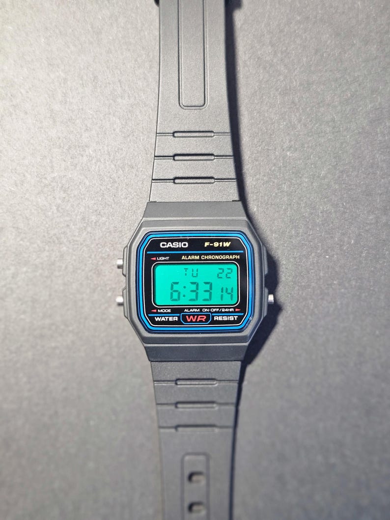May include: A gray Casio F-91W digital watch with a black strap. The watch face has a blue border and displays the time, day, and date. The watch has the text "CASIO F-91W" and "WATER RESIST".