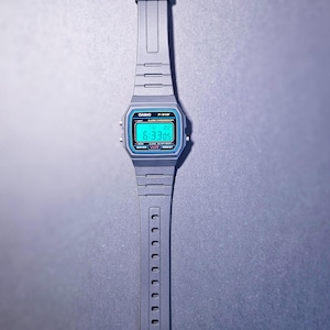 May include: A gray Casio F-91W digital watch with a black strap. The watch face displays the time 6:3305 in a teal-colored font, with a blue border. The watch is on a gray surface.