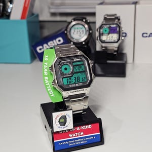 May include: A silver Casio digital watch with a green and black display, showing the time 10:38:12. The watch has a green 10-year battery tag and is displayed on a black stand with "ONE-OF-A-KIND WATCH" text. Other Casio watches are visible in the background.