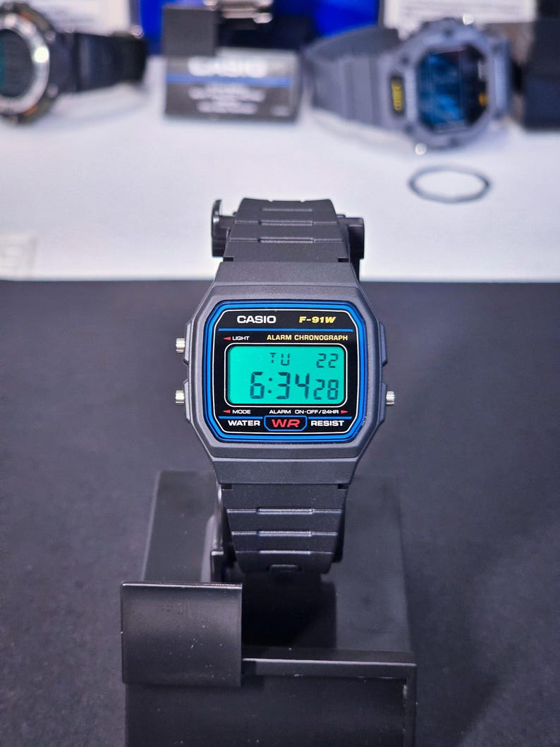 May include: Black Casio F-91W digital watch with a rectangular face and blue accents. The watch displays the time, day, and date. Features include a light, alarm, and water resistance. The watch is displayed on a black stand.
