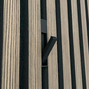May include: A black metal hook fixed to a wall with vertical black and beige striped panels. The hook projects outwards. The wall panels have a wood grain texture. The panels are made of wood.
