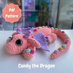 Puede incluir: Un juguete de dragón de ganchillo rosa hecho a mano con alas y detalles morados. El dragón tiene ojos grandes y brillantes y una larga cola. El texto "Candy the Dragon" está en la parte inferior de la imagen y "Pdf Pattern" en un círculo en la parte superior.