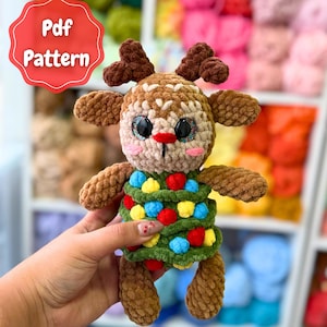 May include: A handmade crochet Christmas Tree Reindeer. The plush toy has brown antlers, a tan face, and a green body decorated with colourful pom-poms. The image includes the text "Pdf Pattern" and "Christmas Tree Reindeer".