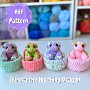Può includere: Quattro giocattoli di drago all'uncinetto dai colori pastello, ciascuno in un cestino abbinato. I draghi sono viola, verdi e rosa, con il testo "Aurora the Hatching Dragon" sotto. Un'etichetta "Pdf Pattern" è sullo sfondo.