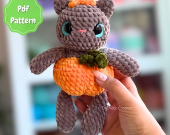 May include: A gray crocheted cat plush toy with large blue eyes, a small pink stitched nose, and a brown stem on its head. The cat is holding a bright orange pumpkin with a green leaf. A green scalloped badge displays the text Pdf Pattern.