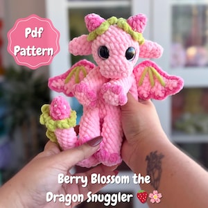 May include: A pink crocheted dragon toy with large black eyes, wings, and a green and pink floral design. The text "Pdf Pattern" is in a pink circle. The text "Berry Blossom the Dragon Snuggler" is below the dragon.