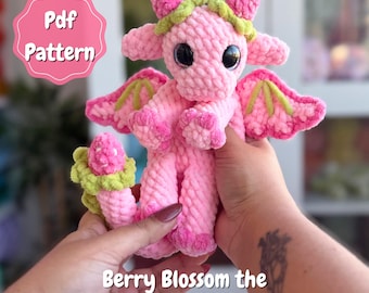 May include: A pink crocheted dragon toy with large black eyes, wings, and a green and pink floral design. The text "Pdf Pattern" is in a pink circle. The text "Berry Blossom the Dragon Snuggler" is below the dragon.