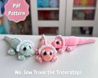 May include: Three crocheted triceratops toys in light green, peach, and pink with white accents. Each dinosaur features large dark eyes and a frill. Text on the image reads: Pdf Pattern and No-Sew Trixie the Triceratops.