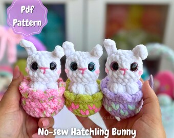 May include: Three crocheted white bunny figures with large eyes, each nestled in a colorful basket. The baskets are pink, green, and purple. The image includes the text "Pdf Pattern" and "No-sew Hatchling Bunny".