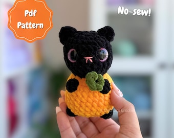 May include: A small, plush, crocheted cat toy with a black head, large purple eyes, and a pink stitched nose. The body is orange with black paws and a small green circular accent. Text on the image reads Pdf Pattern and No-sew!