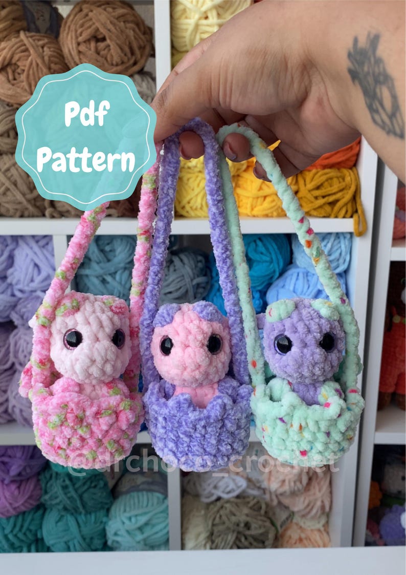 May include: Three handmade crochet dolls in pastel colors, each in a small carrier. The dolls are pink, purple, and mint green, with black eyes. The image includes the text "Pdf Pattern" in a teal circle.