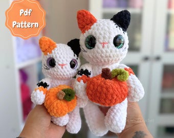 May include: Two handmade crochet calico cats with white, orange, and black yarn, large dark eyes, and pink stitched noses. Each cat holds a small orange crocheted pumpkin with a green stem. An orange badge in the corner reads Pdf Pattern.