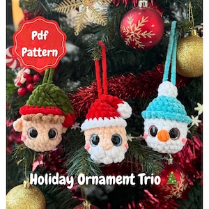 May include: Three handmade crochet Christmas ornaments: an elf, Santa, and a snowman. The elf has a green hat, Santa a red hat, and the snowman a blue hat. Each has large eyes. The text "Holiday Ornament Trio" is displayed. Ornaments are hanging on a tree.
