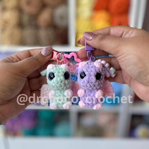 May include: Two crocheted dragon keychains, one mint green and one lavender, are held up. Each dragon has large black eyes, small wings, and a keychain clip. The text "@drofotocrochet" is visible in the image.