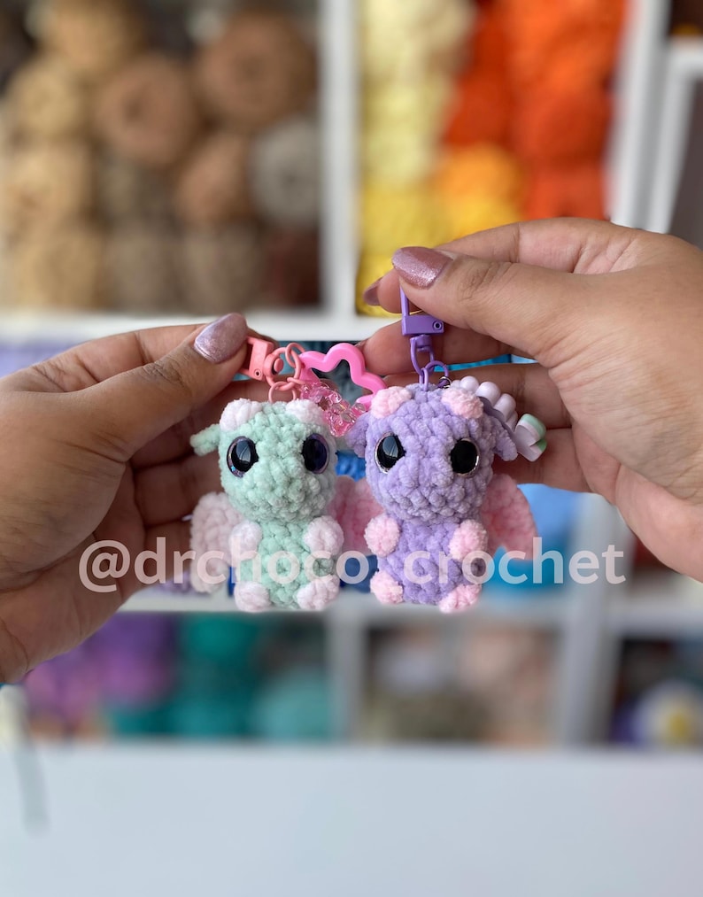 May include: Two crocheted dragon keychains, one mint green and one lavender, are held up. Each dragon has large black eyes, small wings, and a keychain clip. The text "@drofotocrochet" is visible in the image.