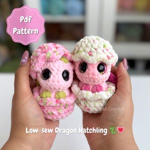 May include: Two handmade crochet dragon hatchlings in pink and white. One is in an eggshell. The dragons have large, sparkly eyes. The image includes the text "Pdf Pattern" and "Low-sew Dragon Hatchling".