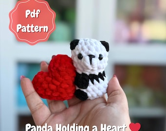May include: A crocheted panda bear with black and white details holds a red heart. The image includes the text "Pdf Pattern" and "Panda Holding a Heart". The panda has black ears and a black patch on its chest.