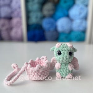 May include: A crocheted dragon and a small pink basket. The dragon is mint green with pink accents and large black eyes. The basket is light pink with a ruffled edge and a handle. The text "@dchoco_crochet" is visible.