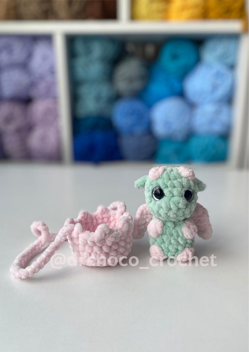 May include: A crocheted dragon and a small pink basket. The dragon is mint green with pink accents and large black eyes. The basket is light pink with a ruffled edge and a handle. The text "@dchoco_crochet" is visible.