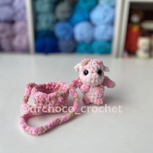 May include: A handmade pink crochet cow with green floral accents and a matching purse. The cow has small wings and a floral crown. The purse has a handle and is also decorated with green floral accents. The text "@archoco_crochet" is visible.
