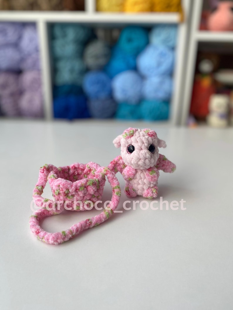 May include: A handmade pink crochet cow with green floral accents and a matching purse. The cow has small wings and a floral crown. The purse has a handle and is also decorated with green floral accents. The text "@archoco_crochet" is visible.