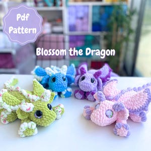 Könnte beinhalten: Vier gehäkelte Drachenspielzeuge in Grün, Blau, Lila und Rosa. Jeder Drache hat große, runde Augen und detaillierte Flügel. Der Text "Pdf Pattern" und "Blossom the Dragon" ist im Bild zu sehen.