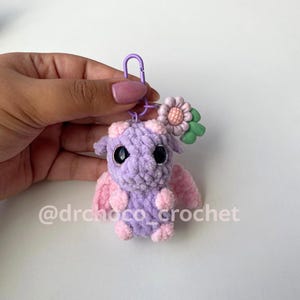 May include: A handmade crochet dragon keychain in shades of purple and pink. The dragon has large black eyes, small pink wings, and a flower accent. The keychain is attached to a purple clasp. The text "@drchoco_crochet" is visible.