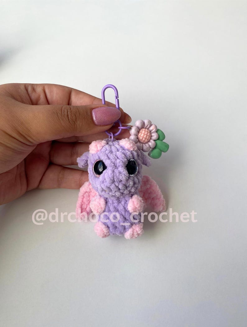 May include: A handmade crochet dragon keychain in shades of purple and pink. The dragon has large black eyes, small pink wings, and a flower accent. The keychain is attached to a purple clasp. The text "@drchoco_crochet" is visible.