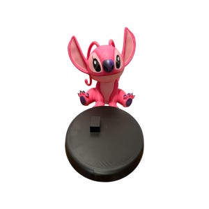 Puede incluir: Figura rosa del personaje Angel de la pel&iacute;cula de Disney Lilo & Stitch. La figura tiene orejas grandes, una nariz morada y est&aacute; colocada sobre una base circular negra. Es un objeto de colecci&oacute;n.