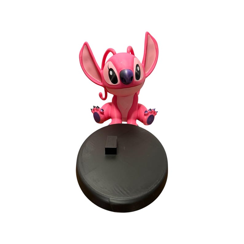 Puede incluir: Figura rosa del personaje Angel de la pel&iacute;cula de Disney Lilo & Stitch. La figura tiene orejas grandes, una nariz morada y est&aacute; colocada sobre una base circular negra. Es un objeto de colecci&oacute;n.
