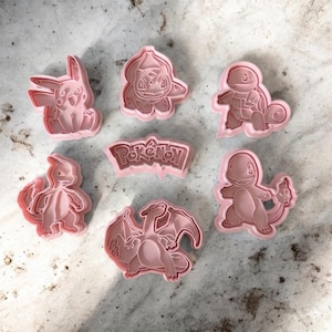 Puede incluir: Un juego de siete cortadores de galletas de plástico rosa claro con varias formas de personajes y una placa central con el texto Pokémon. La colección incluye los contornos de Pikachu, Bulbasaur, Squirtle, Charmander y Charizard.