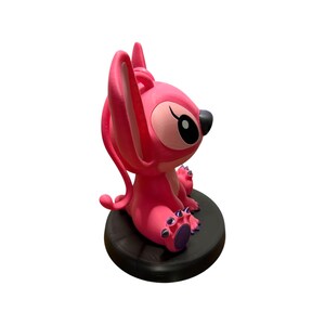 Puede incluir: Figura rosa de Angel de Lilo & Stitch, sentada sobre una base circular negra. La figura tiene orejas grandes, una nariz negra y detalles morados en los pies. Un art&iacute;culo de colecci&oacute;n.