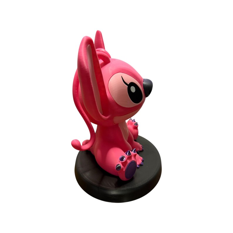 Puede incluir: Figura rosa de Angel de Lilo & Stitch, sentada sobre una base circular negra. La figura tiene orejas grandes, una nariz negra y detalles morados en los pies. Un art&iacute;culo de colecci&oacute;n.