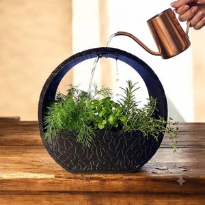 May include: A circular black planter with a textured exterior surface containing various green herbs including dill, basil, and rosemary. A copper-colored watering can pours water into the center of the planter, creating a stream of water.