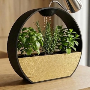 May include: A modern, circular planter with a black frame and a gold-colored textured base. The planter holds various green herbs and is being watered from a silver watering can. The design is contemporary and suitable for indoor use.