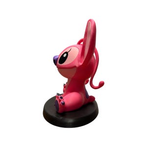 Puede incluir: Una figura rosa de un personaje de dibujos animados con orejas grandes, una nariz morada y detalles morados en los pies. La figura est&aacute; sentada sobre una base circular negra. El personaje est&aacute; en una pose relajada.