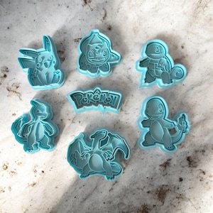 Może przedstawiać: Zestaw jasnoniebieskich plastikowych foremek do ciastek przedstawiających różne postacie z Pokémonów, w tym Pikachu, Bulbasaur, Squirtle, Charmander, Charizard i logo Pokémon. Idealne do tematycznych wypieków.