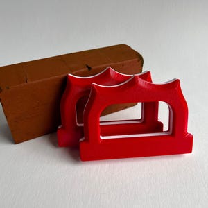 May include: Two bright red, arched-shaped objects with white trim are in the foreground, next to a brown brick. The objects have a decorative top and a rectangular opening. The brick is a reddish-brown color.