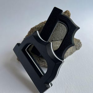 May include: Two black, stylized letter "D" shapes with a decorative, pointed edge design. Each letter has a white accent line. The letters are resting on a rough, gray-brown stone against a white background.