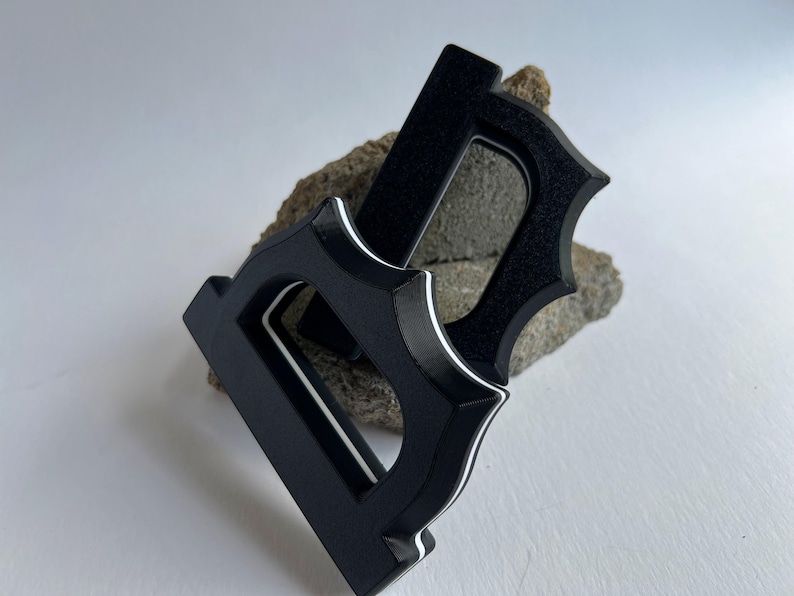 May include: Two black, stylized letter "D" shapes with a decorative, pointed edge design. Each letter has a white accent line. The letters are resting on a rough, gray-brown stone against a white background.