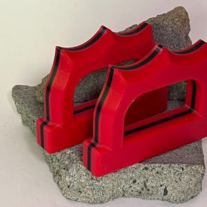 May include: Two bright red, geometric objects with black stripes are displayed on a rough, gray stone. The objects have a unique, arched design with a rectangular opening in the center. The objects appear to be 3D-printed.