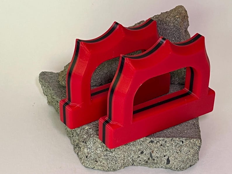 May include: Two bright red, geometric objects with black stripes are displayed on a rough, gray stone. The objects have a unique, arched design with a rectangular opening in the center. The objects appear to be 3D-printed.