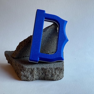 May include: A vibrant blue letter "D" with a white border, resting on a textured gray stone. The letter has a decorative serif design. The background is a plain white surface.