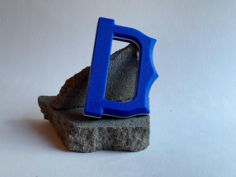 May include: A vibrant blue letter "D" with a white border, resting on a textured gray stone. The letter has a decorative serif design. The background is a plain white surface.