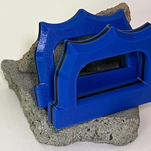 May include: Two blue decorative objects with a black trim, resting on a piece of gray stone. The objects have a unique arched design. The background is a plain white surface.