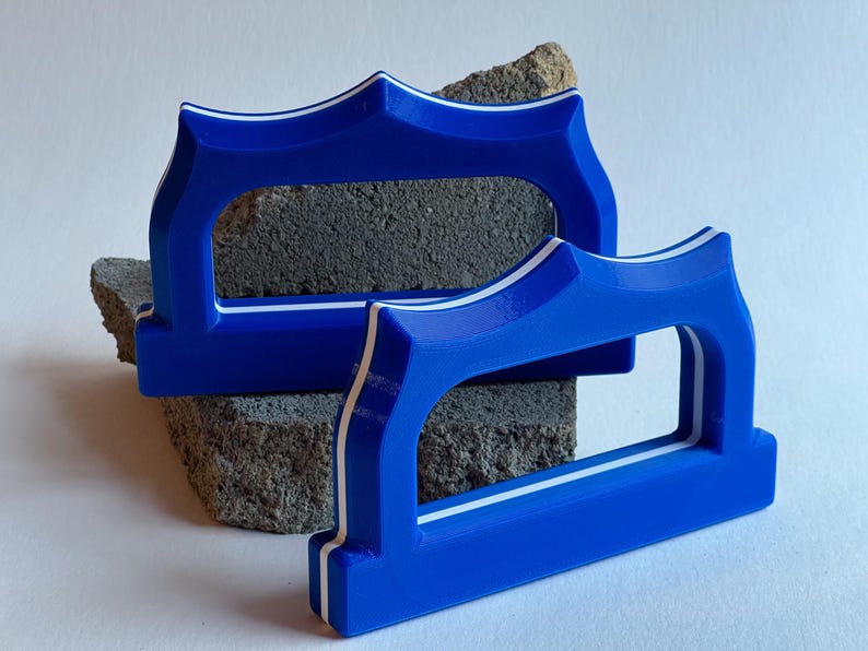 May include: Two blue, shield-shaped handles with white accents. The handles are resting on a rough, gray stone. The handles have a rectangular opening in the center. The handles are likely for a drawer or cabinet.