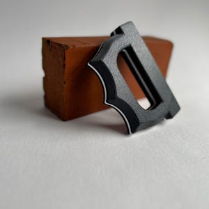 May include: A black metal letter "D" with a decorative design and white trim, resting against a small, reddish-brown brick. The letter has a textured surface and a modern aesthetic, set against a white background.