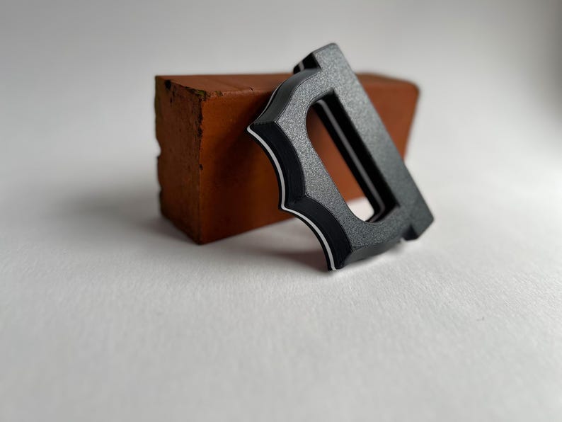 May include: A black metal letter "D" with a decorative design and white trim, resting against a small, reddish-brown brick. The letter has a textured surface and a modern aesthetic, set against a white background.