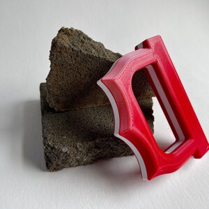 May include: A red and white letter "D" is displayed, leaning against a rough, dark gray stone. The letter is 3D-printed, with a bold, stylized design. The background is a plain white surface.