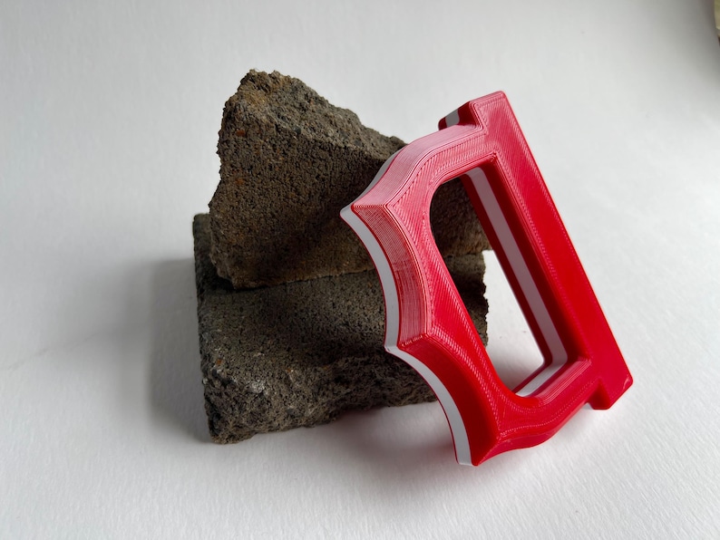 May include: A red and white letter "D" is displayed, leaning against a rough, dark gray stone. The letter is 3D-printed, with a bold, stylized design. The background is a plain white surface.